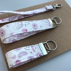 Könnte beinhalten: Drei Schlüsselanhänger aus Stoff mit weißem Hintergrund und einem sich wiederholenden Muster aus rosa und lila Formen. Die Schlüsselanhänger haben silberfarbene Beschläge. Ein Schlüsselanhänger hat eine Handschlaufe und einen silbernen Verschluss. Die Schlüsselanhänger liegen auf braunem kariertem Papier.