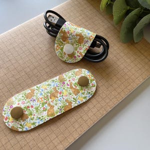 May include: Two fabric cord organizers with a floral pattern featuring small brown rabbits. The organizers have gold metal clasps.