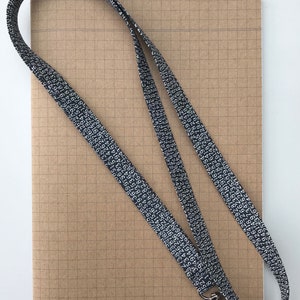 May include: A black lanyard with white numbers printed on it. The lanyard has a silver clip on the end. The lanyard is lying on top of a brown notebook with a grid pattern.