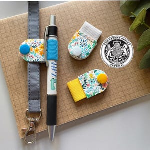 Può includere: Un set di cinque portapenne in tessuto con motivi colorati, tra cui un design floreale blu e giallo, un design floreale bianco, un design floreale giallo e bianco, un design floreale bianco con un bottone blu e un sigillo in bianco e nero con il testo "The Patent Office Official".