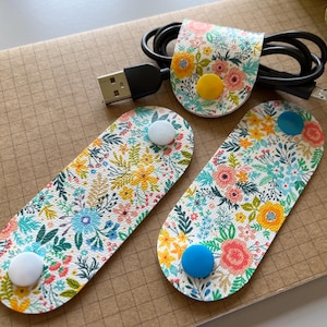 May include: Three floral patterned cord organizers with white, blue, and yellow buttons. The organizers are made of fabric and are designed to hold cords and cables.