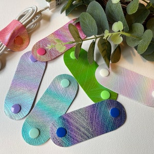 May include: Assortment of colorful iridescent leather cord organizers with different colored buttons. The organizers are designed to keep cords and cables tidy.