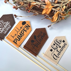 May include: Four small felt flags on wooden sticks with autumn-themed text. The flags are brown, orange, and beige, with phrases like "Hot Chocolate Weather" and "Hello Pumpkin" in white and black script. A fall wreath is in the background.