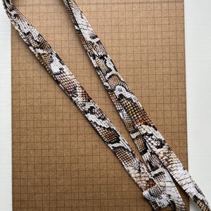 May include: A lanyard featuring a snakeskin design in brown, beige, and white. It has a silver-coloured metal clasp. The background is a brown grid paper.