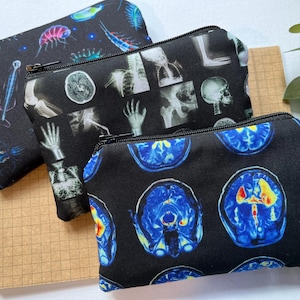 Könnte beinhalten: Drei schwarze Reißverschlusstaschen mit einzigartigen Mustern. Eine zeigt farbenfrohe mikroskopische Organismen, eine andere zeigt Röntgenbilder von Knochen und die dritte blaue und gelbe Gehirnscans. Die Taschen liegen auf einer braunen Papieroberfläche.