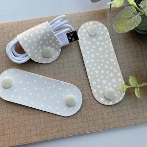 May include: Cable organizers in beige and white with dot and star patterns. One organizer wraps around a white USB cable. Two additional organizers are also shown, each with a snap closure.