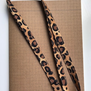 May include: A leopard print lanyard with a silver-colored clasp. The lanyard is tan with brown and black spots. The lanyard is lying on a brown grid paper notebook.