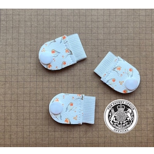 May include: Three white fabric finger protectors with a bird and floral pattern, each with a white button and elasticated band. They are arranged on a brown grid paper background.