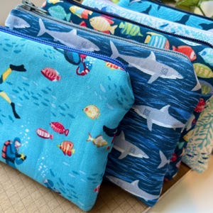 May include: A collection of zippered pouches with aquatic-themed patterns. Designs include scuba divers, sharks, fish, and coral. The pouches are various shades of blue and have blue zippers. These pouches are ideal for storing small items.