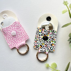 May include: Two small keychains, one pink with white spots, the other with a colourful leopard print. Each has a gold-coloured ring and a snap closure. They are designed to hold small electronic devices. The keychains are approximately 10 cm long.