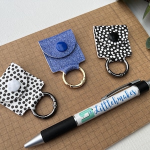May include: Three keychains with different patterns: white with black polka dots, black with white polka dots, and blue denim. Each keychain has a metal ring and a button closure. The keychains are on a brown gridded surface with a pen that says "Littlehmakes".