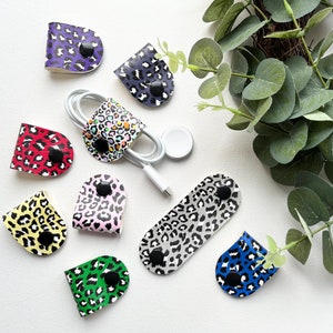 Könnte beinhalten: Eine Sammlung farbenfroher Kabelorganisatoren mit Leopardenmuster und schwarzen Druckknöpfen. Die Organizer sind in verschiedenen Farben erhältlich, darunter Lila, Rot, Gelb, Rosa, Grün, Blau und Grau. Einige Organizer halten weiße Ladekabel und ein weißes Apple Watch Ladegerät.