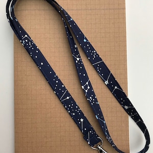 May include: A navy blue lanyard with a white constellation pattern and a silver swivel clasp. The lanyard is lying on a brown gridded surface.