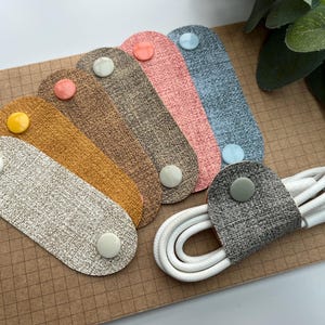 May include: A collection of cable organizers in various colors, including gray, tan, orange, brown, pink, and blue. Each organizer has a button closure. A white USB cable is neatly bundled with a gray organizer.