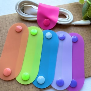 May include: A collection of colorful silicone cable organizers in orange, green, blue, purple, and pink. Each organizer has a matching button closure. A white cable is neatly coiled, held by a pink organizer with a button closure.