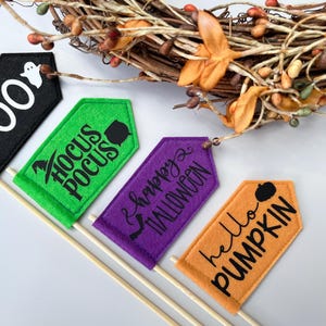 May include: Four Halloween-themed felt tags on wooden sticks. The tags are black, green, purple, and orange, with phrases like "BOO," "Hocus Pocus," "Happy Halloween," and "hello PUMPKIN." A decorative wreath with berries and leaves is in the background.