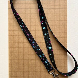 May include: A black lanyard with a silver clip. The lanyard has a repeating pattern of colorful stars on a black background.