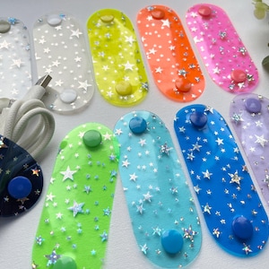 May include: A collection of colorful cable organizers with star patterns. The organizers come in various colors, including clear, yellow, orange, pink, green, blue, and purple. Each organizer has a button closure and is designed to hold and manage cables.