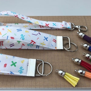 Può includere: Due portachiavi in tessuto bianco con simboli matematici colorati e un anello portachiavi argentato. I portachiavi sono su una superficie a griglia con sei nappe colorate.