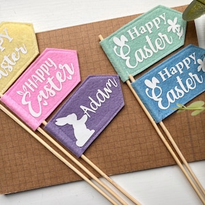 May include: A selection of colourful felt flags on wooden sticks, perfect for Easter. The flags are in shades of yellow, pink, purple, green, and blue. Each flag has the words "Happy Easter" or a name in white script. A white bunny silhouette is on the purple flag.