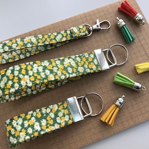 May include: Three keychains with daisy print fabric straps and silver key rings. Each keychain has a different coloured tassel attached: red, green, and yellow.