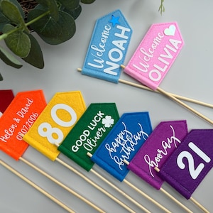 Könnte beinhalten: Eine Sammlung farbenfroher Filzflaggen-Dekorationen auf Holzstäben. Die Flaggen zeigen Zahlen, Namen und Sätze wie "Happy Birthday" und "Good Luck". Farben sind Rot, Orange, Gelb, Grün, Blau, Lila und Rosa. Die Flaggen sind für Feiern gedacht.