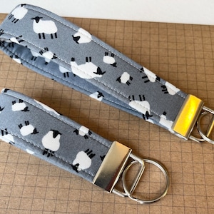 May include: Two grey fabric keychains with white sheep designs and silver key rings. The keychains are different lengths.