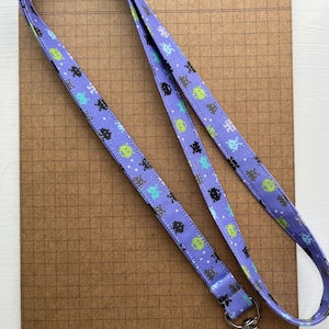 Peut inclure: Un cordon de cou lilas avec un motif répété de personnages extraterrestres pixélisés verts, bleus et noirs. Le cordon a un fermoir en métal argenté et repose sur une surface quadrillée marron.