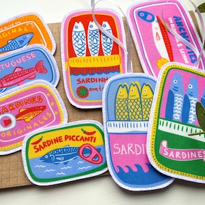 May include: A selection of vibrant fabric patches designed as sardine cans. The patches display fish illustrations and text in multiple languages, such as "Sardines," "Portuguese," and "Originales."