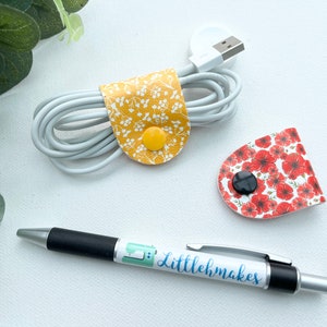 May include: Two fabric cord organizers with button closures. One is yellow with white floral print and the other is white with red poppy print. A pen with the text "littlehmakes" is also pictured.