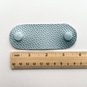 Peut inclure: Une sangle en cuir bleu clair avec deux boutons blancs. La sangle mesure environ 10 cm de long et est mesur&eacute;e &agrave; l'aide d'une r&egrave;gle.