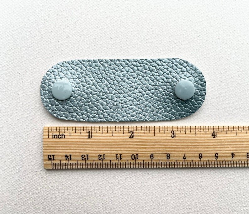 Peut inclure: Une sangle en cuir bleu clair avec deux boutons blancs. La sangle mesure environ 10 cm de long et est mesur&eacute;e &agrave; l'aide d'une r&egrave;gle.