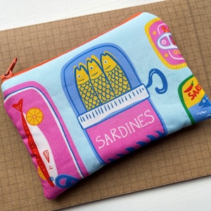 May include: A rectangular, light blue zipped pouch with a colourful sardine design. The pouch features illustrations of sardines in a can, fish, and the word "Sardines" in pink. The zip is orange.