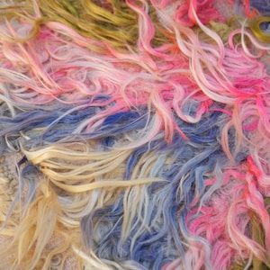 May include: A shaggy textile featuring long, flowing fibers in a multi-colored palette of vibrant pink, soft cream, muted olive green, and dusty blue. The strands are layered and intertwined, creating a dense, textured surface with a wavy appearance.
