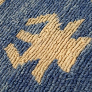 May include: A close-up view of a textile surface featuring a thick, blue, ribbed yarn background with a prominent, cream-colored, geometric, cross-like pattern woven into the fabric. The texture is dense, showing individual yarn strands.