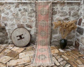 Puede incluir: Una alfombra larga y estrecha con fondo rosa y intrincados patrones florales en tonos beige y crema. La alfombra está colgada en una pared de piedra.