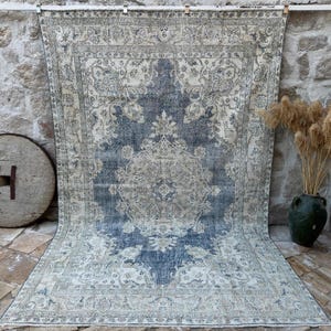 May include: A large, rectangular rug with a central medallion design in shades of blue and cream. The rug has an aged, distressed appearance, with intricate floral patterns throughout. The rug is displayed against a stone wall, with a wooden wheel and a vase of dried plants nearby.