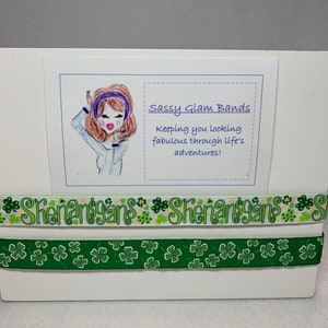 Può includere: Un biglietto bianco con un'illustrazione di cartone animato di una donna con i capelli castani e una fascia blu. Il testo sul biglietto recita "Sassy Glam Bands Keeping you looking fabulous through life's adventures!" Due nastri verdi e bianchi con il testo "Shenanigans" sono sotto il biglietto.