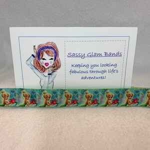 Może przedstawiać: Kartka z tekstem "Sassy Glam Bands" i "Keeping you looking fabulous through life's adventures!"  Kreskówkowa ilustracja kobiety z brązowymi włosami, która nosi niebieską opaskę na głowę i białą koszulę. Zielono-żółta wstążka z kreskówkowym nadrukiem postaci.