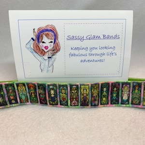 Könnte beinhalten: Eine Karte mit einer Cartoon-Illustration einer Frau, die ein Stirnband und ein blaues Hemd trägt. Auf der Karte steht "Sassy Glam Bands" und "Keeping you looking fabulous through life's adventures!" Vor der Karte liegt ein buntes Band mit einem Buntglasmuster.