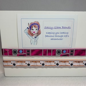 May include: A white sign with a cartoon illustration of a woman wearing a blue headband and a white shirt. The sign says "Sassy Glam Bands" and "Keeping you looking fabulous through life's adventures!" There are two strips of patterned ribbon below the sign.