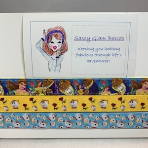 Könnte beinhalten: Drei gelbe, blaue und gelbe Haarband-Haargummis mit einem Beauty and the Beast-Motiv. Die Haarband-Haargummis zeigen ein sich wiederholendes Muster von Belle, Beast und einer Teetasse. Die Karte über den Haarband-Haargummis lautet "Sassy Glam Bands Keeping you looking fabulous through life's adventures!"