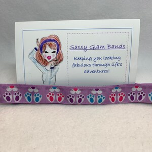 Könnte beinhalten: Lilafarbener Haargummi mit weißem Hintergrund und einem sich wiederholenden Muster aus Pfotenabdrücken in Rosa, Blau und Lila. Der Haargummi wird von einer Karte mit der Aufschrift "Sassy Glam Bands" und "Keeping you looking fabulous through life's adventures!" begleitet.