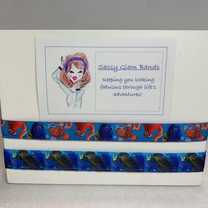 May include: A white display board with two blue and orange ribbon hair ties. The ribbon hair ties feature a Finding Nemo theme with images of Dory, Nemo, and sea turtles. The board also has a white card with the text "Sassy Glam Bands keeping you looking fabulous through life's adventures!" and a cartoon illustration of a woman with a headband.