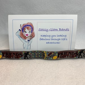Puede incluir: Una pulsera de tela gris con el texto "Friends Don't Lie" en rojo y amarillo. La pulsera también presenta una ilustración en blanco y negro de un monstruo y un círculo amarillo con el texto "Promise?" en negro. La pulsera está sobre un fondo blanco con una tarjeta que dice "Sassy Glam Bands" y "Keeping you looking fabulous through life's adventures!"