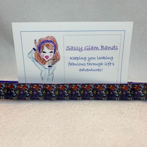Puede incluir: Una liga para el pelo de color morado y negro con un patrón de calavera y flores. La liga para el pelo se muestra en una tarjeta blanca con el texto "Sassy Glam Bands" y "Keeping you looking fabulous through life's adventures!"