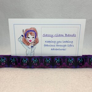 May include: A purple and black ribbon with a skull pattern. The ribbon is sitting on top of a white card with the text "Sassy Glam Bands keeping you looking fabulous through life's adventures!"
