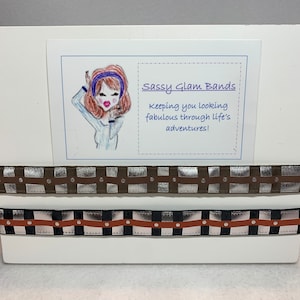 Könnte beinhalten: Ein weißes Präsentationsbrett mit dem Text "Sassy Glam Bands" und dem Slogan "Keeping you looking fabulous through life's adventures!" Zwei braune und silberne Haargummis mit Muster sind auf dem Brett ausgestellt.
