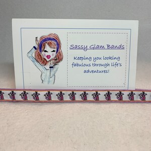 May include: A white card with a cartoon illustration of a woman with brown hair wearing a blue headband. The card says "Sassy Glam Bands" and "Keeping you looking fabulous through life's adventures!" A white ribbon with a pink and orange dragon pattern is below the card.