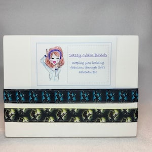 Könnte beinhalten: Zwei Haargummis mit einem schwarzen und blauen Muster und einem schwarzen und grünen Muster. Die Haargummis werden auf einer weißen Oberfläche mit einem Schild präsentiert, auf dem "Sassy Glam Bands keeping you looking fabulous through life's adventures!" steht, und einer Cartoon-Illustration einer Frau mit braunen Haaren, die ein blaues Stirnband trägt.